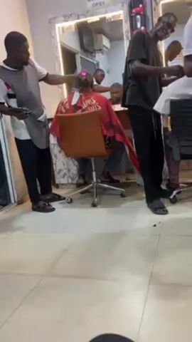 The video shows the interior of a busy barbershop. Four barbers are visible, each attending to a customer in a barber chair. The barbers are all dressed casually in clothes such as t-shirts, trousers, and vests. Some wear aprons. They are using electric clippers to cut the hair of their customers, who are seated with barber capes on. The shop has mirrors on the walls, showing reflections of the activity within. The floor is tiled and a sign reading "OPAY" is visible. The barbers are moving around and interacting with their clients.