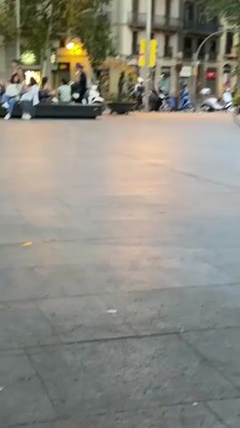 A group of people are sitting on a bench. A man on a bicycle rides by. Another man, shirtless, runs past the bicycle. A couple walks by the bench. People are seen sitting and standing in the background. The scene appears to be an outdoor public space with buildings and trees.
