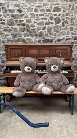 Two large brown teddy bears are sitting on a wooden bench in front of an upright piano. The piano has dark wood and a black and white keyboard. Behind the piano is a wall constructed of irregular gray stones. A blue metal object, possibly a tool, lies on the concrete ground in front of the bench. The teddy bears are positioned side-by-side, facing forward. The bear on the left is slightly closer to the camera.