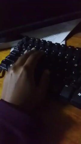 A hand types on a black keyboard on a wooden surface. A piece of paper and a computer monitor sit behind the keyboard. The lighting is dim.