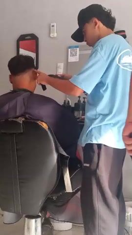 A person wearing a black baseball cap and a light blue t-shirt is using hair clippers to cut the hair of another person sitting in a barber chair. The person in the chair is wearing a dark cape over their shoulders. The barber is holding the clippers in their right hand and appears to be focusing on the back of the seated person's head, near the ear. The environment seems to be a barbershop, with a mirror on the wall behind the barber and various bottles or containers on a shelf in the background. A QR code is visible on the wall.