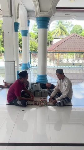 Three men are sitting on a tiled floor in an open-air covered area, possibly a mosque or temple, with pillars supporting the roof. One man on the left, wearing a red shirt and a black skullcap, is interacting with a pile of money. In front of him are several small grey boxes. A second man on the right, wearing a white long-sleeved shirt and a grey skullcap, is also looking at the money. He is wearing striped pants. A third man is lying down behind the pile of money, resting. The floor is white with dark grey lines. In the background, there are trees and a building with a brown roof. The light suggests it is daytime.