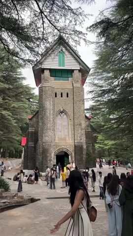 The video captures a full shot of St. John in the Wilderness Church in McLeod Ganj, India, during daylight. The church, constructed from gray stone, features a tall tower, arched windows, and a green accent at the top. In front of the church, a stone-paved area is populated with people, many of whom are dressed in casual attire and interacting in small groups. Some individuals are standing and talking, while others are sitting, suggesting a relaxed atmosphere. In the foreground, a woman with long, dark hair, wearing a light-colored dress, walks towards the left side of the frame. Trees surround the church, partially obscuring the sky and providing a natural frame for the scene.