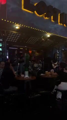 The video depicts a lively scene inside a bar or club, likely in Thủ Đức, Vietnam. A live band is performing on a stage illuminated by bright stage lights. The band consists of at least three members: a guitarist on the right, a bassist on the left, and a drummer in the back. A female singer is actively engaging with the audience, moving around the stage.

In the foreground and mid-ground, patrons are seated at tables, enjoying the performance. Some are facing the stage, while others are engaged in conversation. A prominent green beer tower, with the Heineken logo visible, stands on one of the tables, suggesting a focus on beverages. The lighting is dim overall, with spotlights on the stage creating a dramatic effect.

The atmosphere appears energetic and social, typical of a nighttime entertainment venue. The overall mood seems to be one of enjoyment and engagement with the music. Visible text includes the "Heineken" logo on the beer tower. Above the stage, illuminated signage with yellow lettering against a dark, cloudy background spells out "LOTUS," providing a potential clue to the establishment's name. The time of day is clearly night, and the setting is indoors.