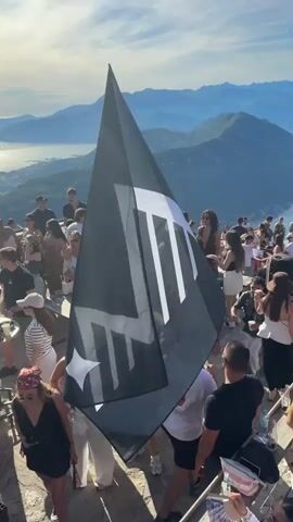 A vertical, handheld video captures a crowded outdoor event on a mountaintop with a panoramic view of a large lake and surrounding mountains. The sky is clear and blue with some clouds. In the foreground, a large black flag with a white logo is being held up. The flag has a stylized white design that includes two distinct shapes, one resembling a star or an asterisk.

A diverse group of people, mostly young adults, are gathered around the flag and enjoying themselves. They are dressed in casual summer attire, including t-shirts, shorts, dresses, and hats. Some individuals are holding drinks. The atmosphere appears festive, with many people looking towards the camera or interacting with each other.

The ground beneath the people is rocky and uneven, suggesting a natural, elevated location. Several tables and chairs are visible, indicating a setup for an event or gathering. The background reveals the expansive vista of the lake, which is dotted with boats, and the layered mountain ranges stretching into the distance under the bright sunlight. The overall impression is one of a lively celebration or party taking place in a scenic, high-altitude setting.