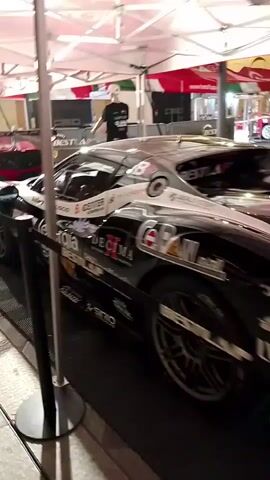 The video shows a pit area with several racing cars parked under canopies. The camera pans from right to left, focusing on a black racing car with various sponsor logos and the number 8 visible. In the background, a red racing car with a large spoiler is also visible. Several people are present in the pit area, some wearing racing attire. The environment appears to be an outdoor event, possibly a race track, with tents and lighting. The overall impression is of a busy pre-race or post-race setup.