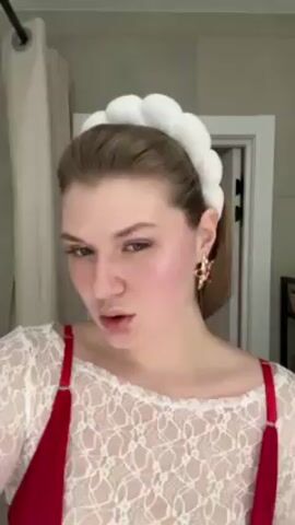 A young woman with fair skin and light brown hair pulled back into a low ponytail poses in front of a bathroom mirror. She is wearing a white lace top over a red garment, a white cloud-like headband, and large gold earrings with an eye design. The bathroom has a white shower curtain and a white door in the background. The woman makes a kissy face, then opens her mouth in an exaggerated O shape. She then touches her neck and rests her chin on her hand, looking away. The video is accompanied by upbeat music.