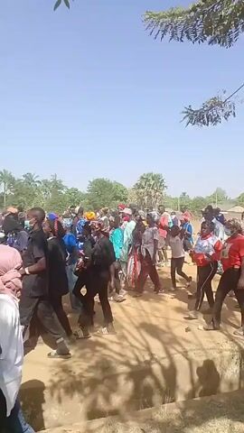 The video shows a crowd of people walking along a dirt path under a clear blue sky.  The landscape includes trees and hills in the background.  The individuals are dressed in a variety of casual clothing, with some wearing colorful head coverings and hats.  The group appears to be moving together, possibly as part of a procession or walk.  Some people are carrying items, and the general atmosphere seems relaxed and communal.
