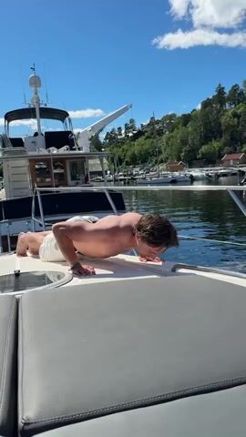 A young, shirtless man is performing push-ups on the deck of a boat. He is wearing white shorts. The boat is in a harbor, with other boats docked behind it. In the background, there is a lush green hillside with houses and trees. The sky is a clear blue with a few white clouds. The sun is shining brightly, indicating it is daytime, likely midday or early afternoon. The water is calm and reflects the blue sky. The overall scene suggests a sunny day in a scenic, possibly Scandinavian, location, given the "Asker, Norway" context. The man appears focused on his exercise. There is no visible text in the media.