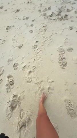 The video begins with a close-up view of a person's bare feet walking on a sandy beach, with footprints visible in the sand. The camera then pans out to reveal a wider view of the beach, with the ocean and a cloudy sky in the background. A distant island and buildings can be seen along the coastline. The waves gently lap at the shore, and the overall atmosphere is calm and serene. Several other footprints are scattered across the sand, suggesting that other people have been on the beach. In the distance, a few figures can be seen walking along the shoreline and near the buildings. The sky is filled with dramatic clouds, with hints of blue sky peeking through in some areas. The camera continues to pan, offering a panoramic view of the beach and the ocean.