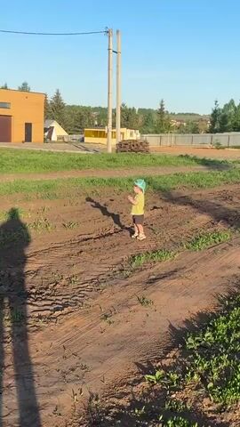 The video shows a young child standing in a muddy patch of land. The child wears a light green t-shirt, dark shorts, a turquoise baseball cap, and light-colored shoes. The ground is muddy and uneven with sparse green vegetation. In the background, there are various structures, including a yellow building with a white trim, a pile of wooden planks, a fence, and trees. Two utility poles stand near the child. The sunlight casts a distinct shadow of the child on the ground. The child appears to be looking down and seems contemplative.