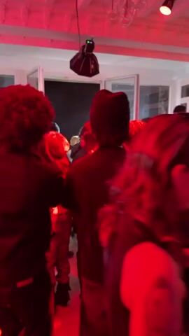 The scene is a crowded indoor space bathed in red light. A group of people stand relatively still, many facing toward open doors at the far end of the room. Several people wear hats or beanies. A lighting fixture hangs from the ceiling. The room appears to be a party or gathering.