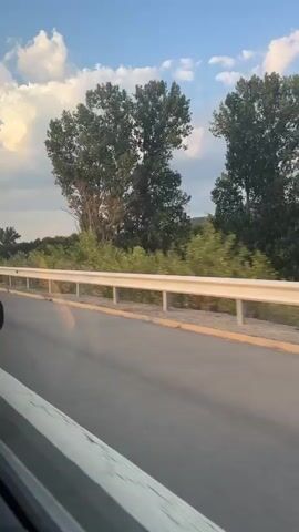 The video shows a view from inside a vehicle, looking out the window at a highway and the surrounding landscape. The road is paved, with a white line marking the lane. A guardrail runs along the side of the road, separating it from a grassy area. Beyond the guardrail, there are trees and vegetation, and in the distance, hills or mountains. The sky is blue with white clouds, some of which are illuminated by sunlight. The perspective suggests the vehicle is in motion, likely traveling along the highway. There are no people visible in the frame. The overall impression is one of a scenic drive through a rural or semi-rural area.