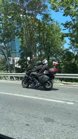 The video shows a black motorcycle with two riders wearing helmets and jackets, traveling on a road with traffic. The motorcycle has saddlebags and a top case. In the background, there are buildings, trees, and a clear blue sky. The camera is inside a vehicle, as indicated by the presence of a side mirror. The motorcycle passes under a bridge with a white railing. There are a few small flags on the back of the motorcycle.