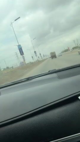 The video captures a first-person perspective from the dashboard of a car driving on a multi-lane road in Lugbe, Nigeria. The scene unfolds on a cloudy or overcast day, with the sky appearing gray and diffused light illuminating the environment.

The foreground is dominated by the car's dashboard, primarily dark gray or black plastic, with the "AIRBAG" label faintly visible. The windshield, seen through, has some small water droplets or smudges. Ahead on the road, a large, dark-colored dump truck, possibly carrying a light-colored load, is prominently visible, moving in the same direction as the filming vehicle. Other cars can be seen further down the road in the distance.

Along the sides of the road, numerous tall streetlights with banner-like attachments are visible, though any text on them is illegible. In the background, what appear to be electricity pylons or large utility poles rise above the landscape, and some sparse vegetation or undeveloped land can be glimpsed. The road itself appears wide and well-maintained, suggesting a major thoroughfare or expressway.

The primary activity is driving, with the car moving forward, following the dump truck. The overall atmosphere is mundane, characteristic of a routine drive. Ambient music, possibly hip-hop, is audible, contributing to a casual feel for the occupants of the car. No specific emotions are discernible from the visual content.