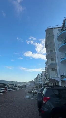 The video pans across a seaside promenade with a row of white apartment buildings on the right and the ocean on the left. The sky is a clear blue with scattered white clouds. A dark-colored SUV is parked in the foreground. The promenade is paved with brick and lined with a white railing. Beyond the railing, the ocean waves are crashing against the shore. There are some small tables and chairs set up along the promenade, likely for outdoor seating at a cafe or restaurant. The buildings have multiple stories with balconies, and some of them have blue accents. The overall atmosphere appears to be that of a coastal town on a windy day.
