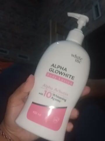 The video shows a close-up of a person's hand holding a white plastic bottle of "ALPHA GLOWWHITE Body Lotion" with a pink label. The bottle has a pump dispenser. The label also indicates it contains "Alpha Arbutin with 10 Whitening Agents" and has a UV protection symbol. The bottle is turned to show the ingredients list and a barcode. The background appears to be indoors with a dark, textured wall.