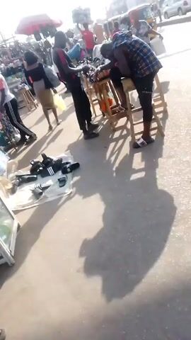 The video shows a busy outdoor market scene, likely during the day, with many people walking around. There are vendors displaying goods for sale on tables and the ground. One vendor is showing slides to a customer who is leaning on a stool.  Another vendor seems to be arranging his merchandise. The surroundings include a street with vehicular traffic. The scene appears to be taken in a crowded, urban environment.
