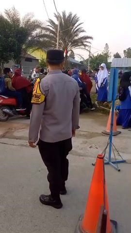 The video shows a group of students, mostly young girls wearing blue uniforms and hijabs, standing outside a building with a green facade. A person in a police uniform with a black cap and a yellow armband is seen walking away from the camera towards the students. Two orange traffic cones are visible in the foreground. The environment appears to be outdoors, possibly in a schoolyard, with trees and a palm tree in the background. The lighting suggests it is late afternoon. Some motorcycles are parked in the background. The students are looking in different directions, and some appear to be talking to each other. One student on the far right is looking at the camera.