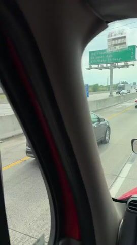 A short video clip shows the interior of a vehicle on a multi-lane highway, with traffic visible through the windshield and side window. The vehicle appears to be a truck, given the size of the mirrors and the visible dashboard. There's a white hardhat on the dashboard, and an "ElitePass" device attached to the windshield near the rearview mirror. A navigation screen displays a route. Another truck, red in color, is visible in the side mirror and then passes by on the left. The highway appears busy, with multiple cars and trucks in both directions. The weather is overcast.
