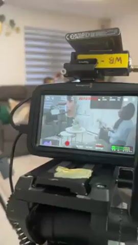 A camera is filming a scene in a living room. The monitor attached to the camera shows two people sitting in chairs, in the middle of the frame. A man stands between them, holding a piece of paper, while gesturing. On top of the monitor is a yellow battery pack with the words "WB -BM" written on it. A person in an orange t-shirt walks in front of the camera. The living room has modern furniture, including a gray couch and a white entertainment console. There are two round decorative tables in the middle of the room. The walls are white, and the lighting is bright.