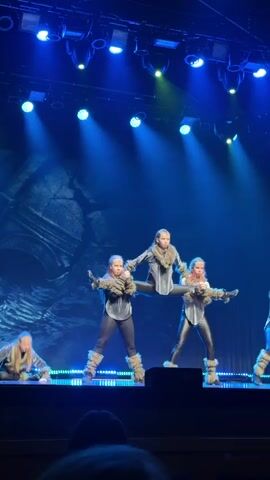 A young woman with a long blonde ponytail, dressed in a grey top, black leggings, and furry leg warmers, is dancing on a stage. She is positioned in the center of the frame, facing slightly right, with her right arm raised and her left hand near her waist. To her immediate right, another young woman in a similar outfit and hairstyle performs a synchronized dance move, mirroring the first woman's posture with her left arm raised. To the left of the central dancer, a third woman in a grey outfit and furry leg warmers is on the stage floor, seemingly performing a push-up. A fourth dancer is visible in the background to the right. The stage is illuminated by blue and green spotlights from above, casting dynamic shadows. The background features a large, dark, rock-like structure. The overall scene depicts a choreographed dance performance.