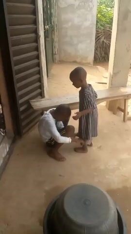 A barefoot child in a striped outfit stands still while another barefoot child in a white shirt and dark shorts crouches down and examines the first child's foot. The crouching child appears to be holding the standing child's foot. They are both outside in what appears to be a yard, with a wooden bench behind them, a door with slats to the left, and a large, dark-colored basin in the lower foreground.