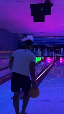 A person in a white t-shirt and black shorts is bowling in a bowling alley. The bowling alley has purple and blue neon lights. The person swings the bowling ball back and then forward to release it down the lane. The ball rolls towards the pins. The person watches the ball as it travels down the lane. The person then walks away from the lane. The American flag is visible on the wall.