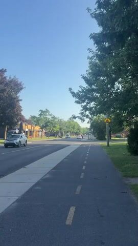 The video shows a view from a bike path running alongside a street. The bike path is asphalt and has dashed yellow lines marking its edge. To the left of the bike path is a sidewalk, then a road with cars, and buildings on the other side. Trees line the street and cast shadows. The sky is clear and blue. A yellow stop sign is visible on a pole on the right side of the bike path. The timecode indicates the video is very short, only showing a few seconds of this scene.