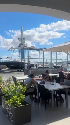 This video captures a sunlit outdoor dining area overlooking a marina. Several people are seated at white tables, some eating or drinking. In the background, a boat is being lifted by a blue crane out of the water. The sky is bright blue with scattered white clouds. Green plants are visible in the foreground in a planter box with a bicycle symbol. The overall atmosphere appears to be a casual daytime scene at a waterfront cafe.