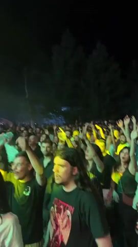 The video shows a crowded outdoor concert at night. The audience is packed together, with many people raising their hands in the air. Stage lighting, primarily yellow and red, illuminates the crowd and the smoke or haze in the air. The background is dark, with trees and possibly tents or structures visible. People are diverse in age and appearance, wearing casual clothing. Some individuals are holding drinks or phones. The overall atmosphere appears to be energetic and engaged with the performance.