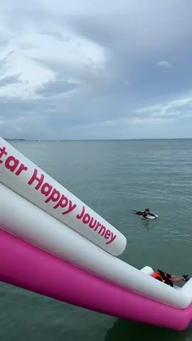 An inflatable slide with "Happy Journey" written on it in red and white is partially visible on the left side of the frame. The slide extends into a body of water. There are several people in the water. One person wearing an orange life vest is splashing as they exit the slide. Another person is on a surfboard in the water. Several other individuals are visible swimming or floating in the water. The sky is cloudy, and the water appears calm.