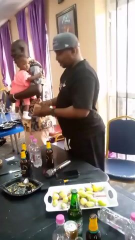 The video depicts a room with two men and a baby. One man is holding the baby, who is wearing a pink outfit, while the other man stands at a table. The table is covered with bottles of beer, water, a microphone, a cell phone, lemon slices, and dirty plates. The room has purple curtains, blue chairs, and a framed picture on the wall. The man at the table is wearing a black shirt, black pants, and a grey baseball cap. The man holding the baby is wearing a patterned shirt and khaki shorts.  It appears to be some kind of social gathering.