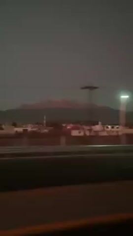 The video shows a mountain range in the distance, viewed from a moving vehicle on a highway. The sky is a dusky grey. In the middle ground, there are buildings, possibly a town or city, with white roofs visible. The camera is likely positioned inside a car, looking out the window as it travels along the road. The road is black, and there's a barrier or wall along the side. A street light is faintly visible on the left side of the frame. The scene has a slightly blurred quality due to the motion of the vehicle.