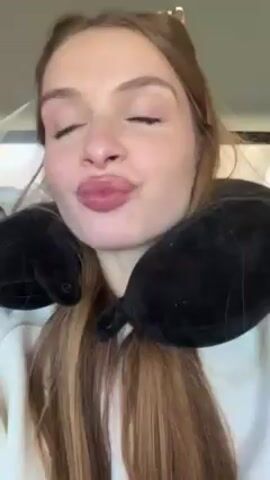 A fair-skinned, young woman with light brown hair and a black travel pillow around her neck sits in a car. She has red nail polish on her nails. She speaks directly into the camera. The interior of the car is visible behind her.