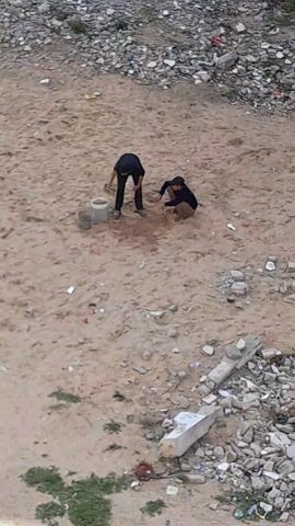Two people are on a sandy ground. One person is standing and pouring something from a small trowel into a bucket. The other person is kneeling and appears to be digging in the sand with their hands. The area is surrounded by debris, including rocks, concrete fragments, and trash.