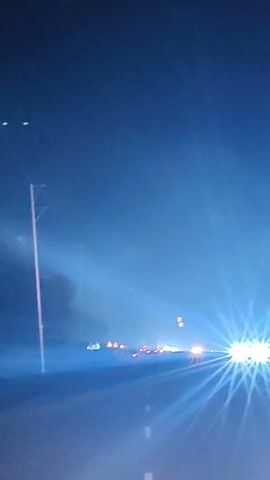 The video captures a nighttime driving perspective on a road, likely a highway, with dense fog or mist obscuring visibility. The headlights of oncoming vehicles create bright, starburst-like flares due to the atmospheric conditions. Several vehicles are visible in the distance, their lights also diffused by the fog. The road surface is dark, with white lane markings intermittently visible. Utility poles line the left side of the road. The sky is a deep, dark blue, consistent with nighttime. There are no people or specific interactions clearly depicted, only the environmental conditions and the act of driving through them.