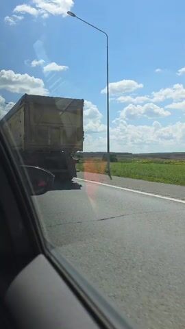 The video captures a rear view of a yellow semi-trailer truck driving on a paved road under a bright blue sky with scattered white clouds. The truck has a large yellow cargo container with some text and a circular logo on its side. The truck has visible red mud flaps behind its rear wheels. The camera appears to be positioned inside another vehicle, looking out the passenger side window, as evidenced by the reflection on the glass and the partial view of the interior of a car. The road is bordered by a green field with some vegetation. The video briefly shows the truck passing and then the view shifts to the open road and fields.
