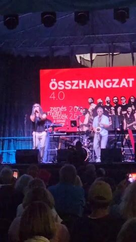 A group of people are performing on a stage in front of a large red screen. On the left, a man with long dark hair is singing into a microphone, wearing a dark t-shirt and light-colored pants. Next to him, a keyboardist is playing a red keyboard. In the center right, a man is playing a saxophone, wearing a light-colored short-sleeved shirt and light-colored pants. Behind them, a drummer is visible. A group of people, presumably the rest of the band, are standing on the stage behind the performers. The red screen displays the word "ÖSSZHANGZAT" in large white letters and "4.0" below it. Smaller text in white is also visible on the screen. The stage is lit, and there are lights above the stage. In the foreground, an audience is seated, with their backs to the camera, some holding up phones to record the performance. The overall setting appears to be an outdoor evening concert.