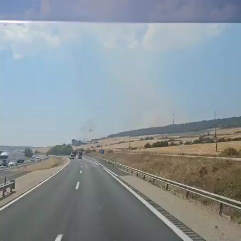 The video shows a view from a vehicle traveling on a highway. The highway has multiple lanes and is bordered by a dry, grassy shoulder and a metal guardrail. The landscape on either side of the highway consists of fields of dry, golden grass, some scattered trees, and rolling hills in the distance. A wind turbine is visible on a hill in the background. There are a few vehicles on the highway, including cars and trucks. In the distance, there is a plume of smoke rising from behind a hill, suggesting a fire or controlled burn. The sky is blue with scattered clouds.
