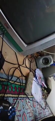 The video shows a close-up view of various electronic devices on a patterned rug. A television set with a green frame is visible in the upper portion of the frame. To the right of the television, a small gray speaker is placed. Several black cables are tangled around the speaker and the television. A beige remote control with many buttons lies on the rug. Two round, colorful objects resembling fans or decorations are also on the rug. A black gaming controller with blue buttons is connected to a black console or base unit. The background consists of wooden walls and a blue plastic bag. The overall scene appears to be a cluttered area with electronic equipment.
