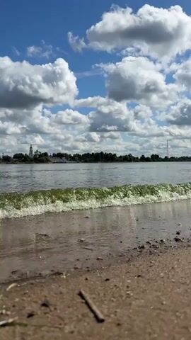 The video shows a sandy beach in the foreground with a body of water in the background. The water is calm with small waves gently lapping at the shore. In the distance, across the water, there is a treeline and a cluster of buildings, including what appears to be a white church with a steeple. The sky is blue with scattered white, puffy clouds. The camera angle is low, looking across the sand and water towards the distant shore. There are no people visible in the video. A small twig lies on the sand in the foreground. The overall impression is a peaceful, natural scene on a sunny day.