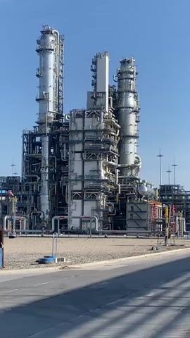 The video shows a wide view of a large industrial facility with multiple tall towers, pipes, and structures under a clear blue sky. The camera pans from left to right, revealing more of the complex. In the foreground, there is a gravelly area and a paved road. There are no people visible in the video. The facility appears to be a chemical plant or refinery. The overall environment is arid with sparse vegetation, suggesting a dry climate.