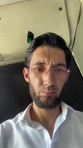 A man with dark hair, a beard, and glasses is shown in a selfie video. He is wearing a white collared shirt. The background appears to be the interior of a vehicle, possibly a bus or train, with a ceiling and a dark surface behind him. The man looks directly at the camera with a neutral or slightly concerned expression.