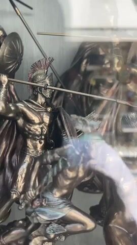 The video shows a display window of a shop, likely an art or souvenir shop, with bronze statues of mythological or historical figures. In the foreground, there are reflections on the glass, obscuring some of the details. The statues depict warriors in armor, some holding spears and shields, and a nude figure entwined with a snake. Behind the statues, shelves are visible, stocked with various items, possibly more statues or decorative objects. The shop appears to be located on a street with a paved sidewalk. The overall scene is somewhat blurry due to reflections and the glass, creating a sense of depth and layering.