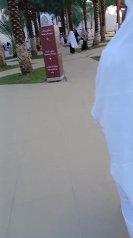 The video shows people walking near a building in an outdoor environment with palm trees. Many of the people are wearing white robes or garments. The ground is paved with light-colored tiles, and there are small patches of green grass. A brown sign with white lettering indicates directions to "Men's Prayer," "Woman's Prayer," and "Bus parking." The camera moves, following a person dressed in white as they walk along the pathway.