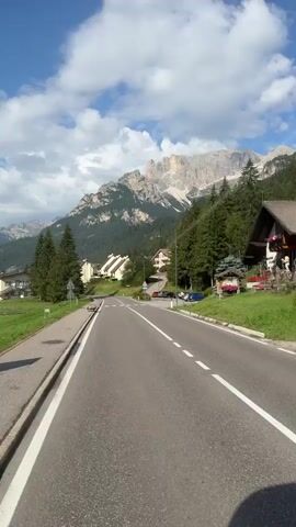 The video shows a paved road with white lines marking the lanes, leading into a mountainous region. The road is flanked by a grassy verge and a sidewalk on the left. On the right side of the road, there are several houses with wooden accents and red roofs, characteristic of a European alpine village. Pine trees cover the slopes of the mountains, which are rocky and rugged, with some snow visible at the higher elevations. The sky is blue with scattered white clouds. A black car is driving away from the camera on the right lane of the road. Further down the road, a white car is also visible. The overall impression is a scenic drive through a mountain landscape with a small village.