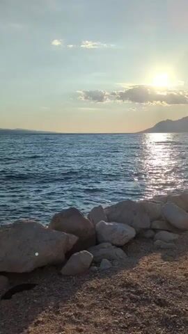 The video captures a serene seascape at sunset. The camera is positioned at eye level, looking out towards the ocean. In the foreground, a rocky shoreline is visible, with large, light-colored boulders scattered along the edge of the water. The ocean itself is a deep blue, with gentle waves reflecting the golden light of the setting sun. The sun is low in the sky, casting a warm glow and creating shimmering reflections on the water's surface. A few wispy clouds are present in the sky, adding depth to the scene. In the distance, a faint outline of landmass can be seen on the horizon. The overall atmosphere is peaceful and tranquil, evoking a sense of calm and natural beauty.