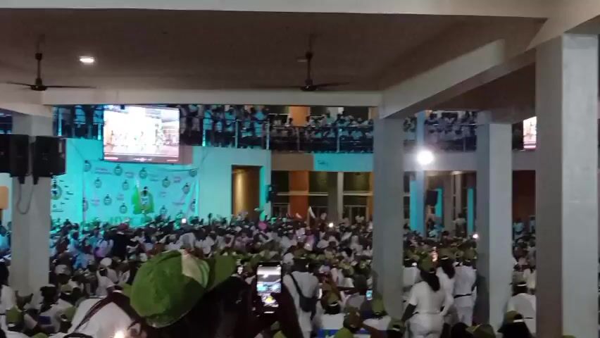 The video shows a large indoor gathering of people, most of whom are wearing white outfits and green hats. The crowd is densely packed, filling the main floor and spilling onto elevated balconies. In the background, there is a large stage area with a banner and a large screen displaying an image. Lights illuminate the stage and parts of the crowd. On the left side of the stage, there are two large black speakers. In the foreground, several people are holding up their mobile phones, seemingly recording the event. The overall atmosphere appears to be energetic and celebratory, with many hands raised in the air. The environment seems to be a hall or convention center, with concrete pillars and a high ceiling.