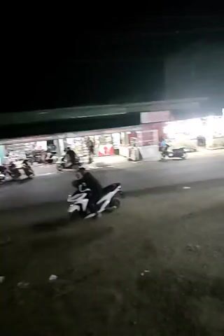 The video shows a nighttime scene on a street with several food stalls or small shops.  Motorcycles are parked and some are in motion.  In the foreground, a person wearing dark clothing is partially visible, leaning on a railing.  Further down the street, illuminated by lights from the stalls, people are visible, and there are more motorcycles and what appear to be tables and chairs set up outside the stalls.  One stall has a display of what looks like coconuts or similar round objects on a stand. The overall atmosphere appears to be a bustling evening market or street-side dining area.